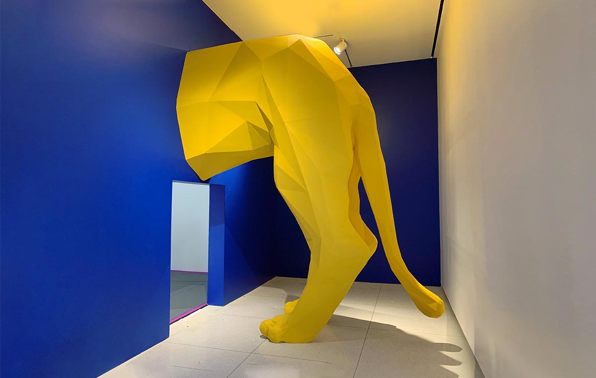 A CNC fabricated bold yellow lion sculpture appears to emerge from a wall at a museum